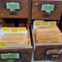 Open wooden card catalog drawers filled with seed packets from the KPU Seed Library, organized by vegetable types such as tomato, kale, cabbage, peppers, and radish.