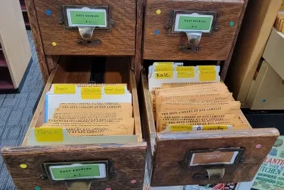 Open wooden card catalog drawers filled with seed packets from the KPU Seed Library, organized by vegetable types such as tomato, kale, cabbage, peppers, and radish.